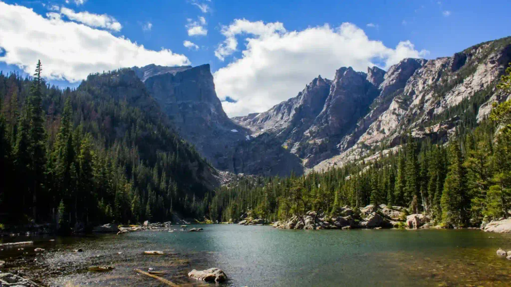 Scenic clear alpine lake surrounded by dense green pine forest and majestic rocky mountain peaks under a cloud-filled blue sky