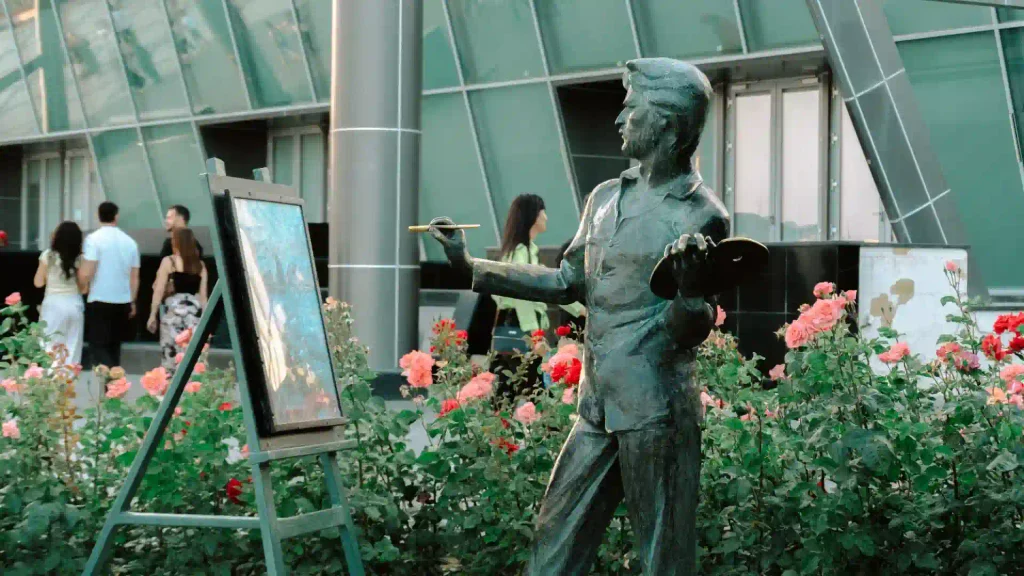 Bronze statue of a painter with a palette and brush next to an easel, surrounded by vibrant blooming pink rose bushes