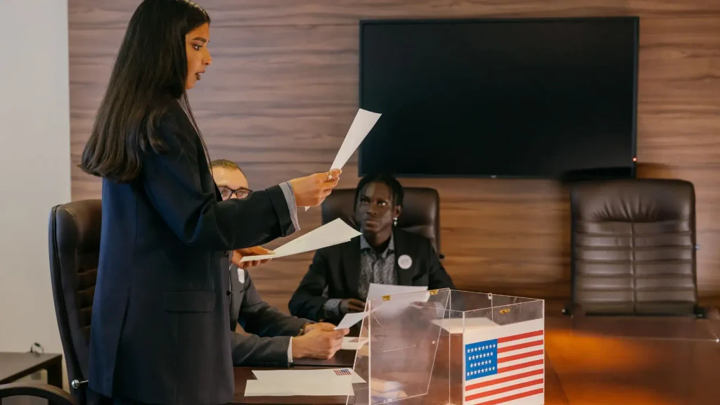 Woman standing and reviewing election paperwork at a board table next to a clear ballot box decorated with an American flag