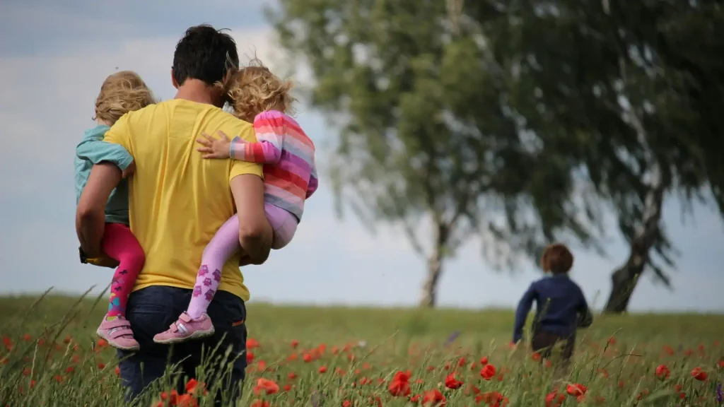 Rear view of a father carrying two toddlers in his arms while walking through a field filled with blooming red poppies