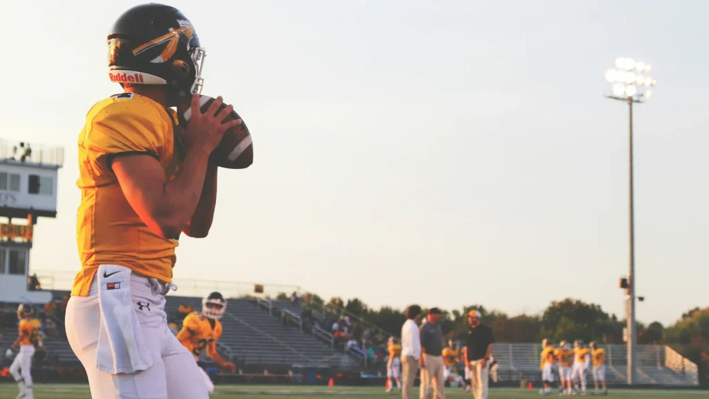 American football quarterback in a yellow jersey holding the ball on the field during practice under bright stadium lights