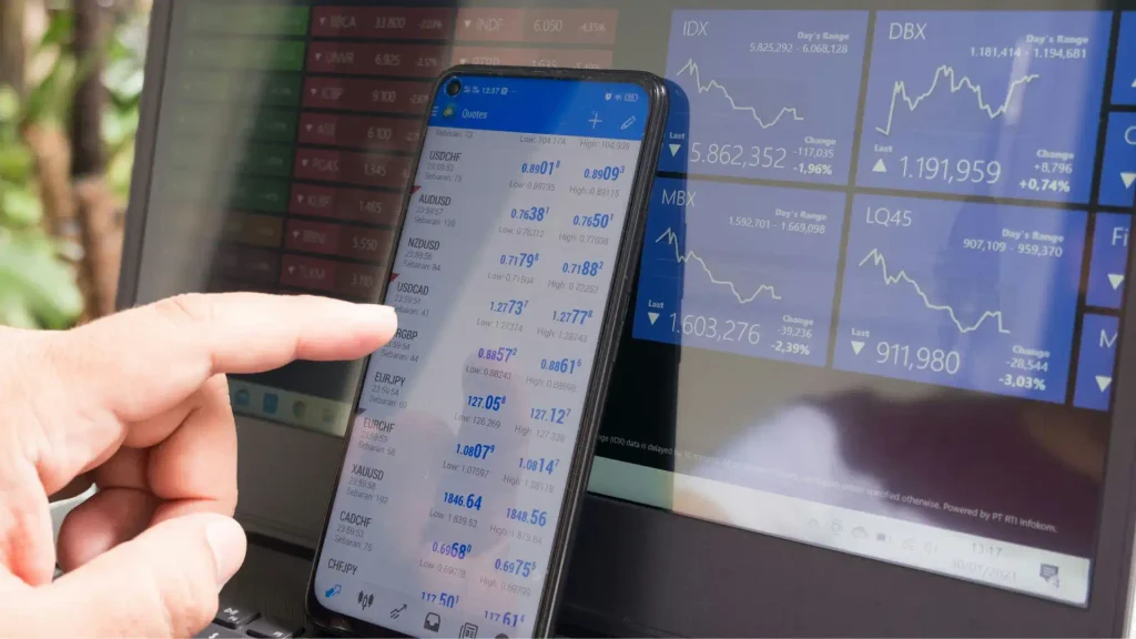 A hand holding a smartphone displaying forex currency trading rates positioned over a laptop displaying real-time stock graphs
