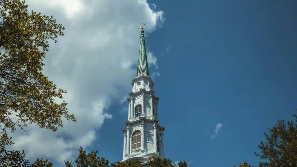 A tall historic white church steeple rising high above lush green tree branches into a blue sky filled with white puffy clouds