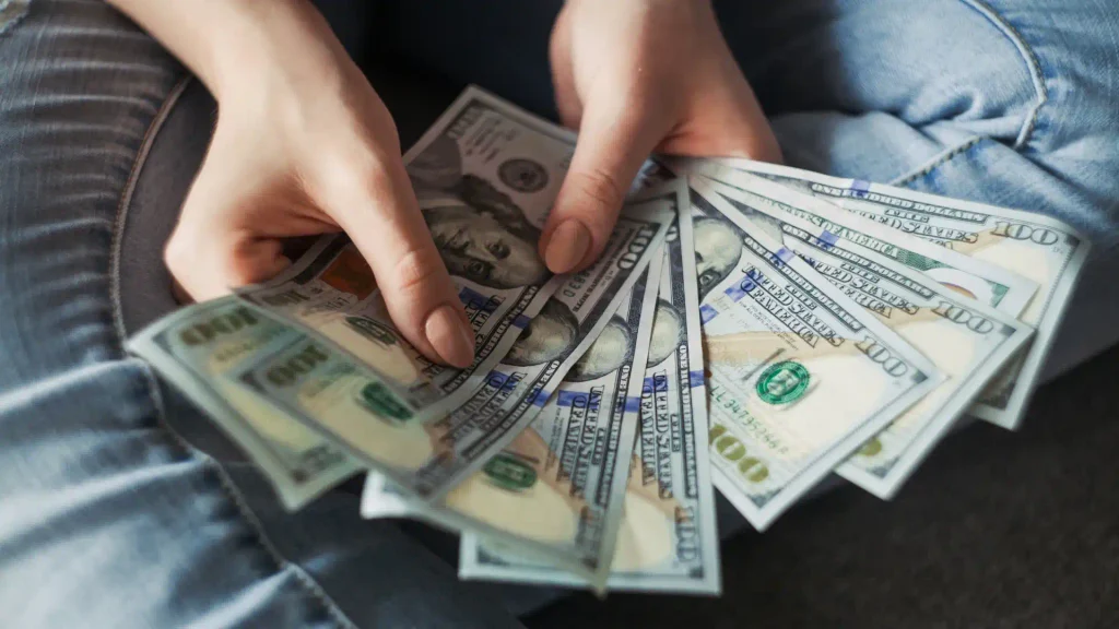 Hands holding and fanning out a stack of hundred-dollar cash bills over denim jeans, showing personal savings or income