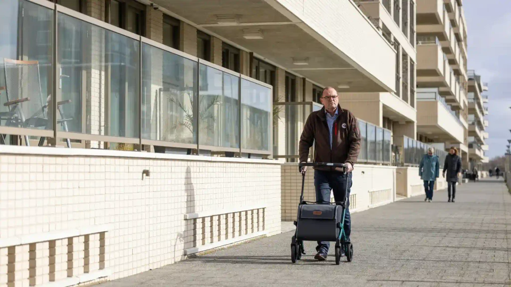 An older man walks along a paved sidewalk past apartment buildings, pushing a dark-blue four-wheeled mobility rollator walker