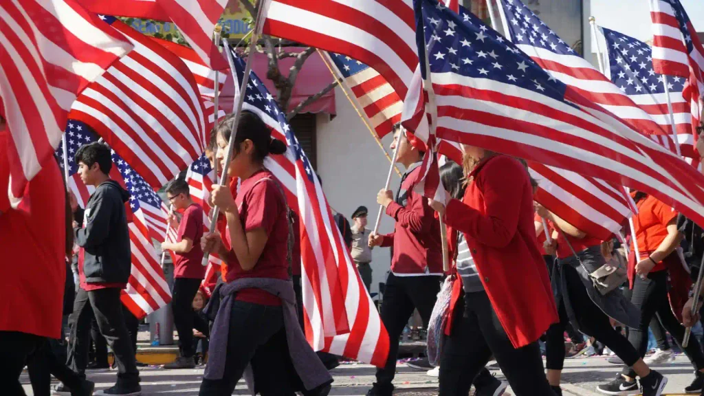 Group of people marching in a patriotic parade while carrying large American flags attached to tall poles on a city street