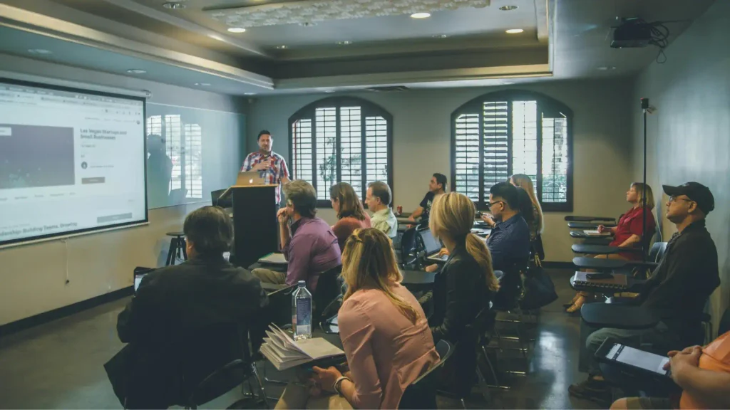 A speaker presenting to a small group of people sitting at desks in a classroom with a large projector screen on the left side