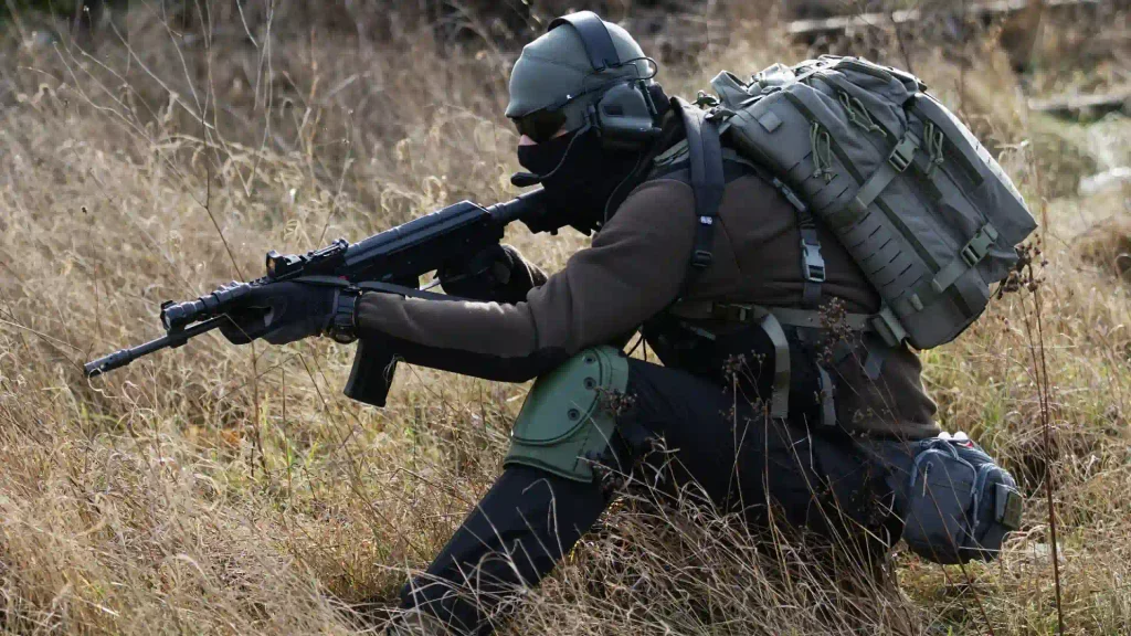 Tactical soldier in full combat gear and balaclava kneeling in tall dry grass while aiming a rifle during field exercises