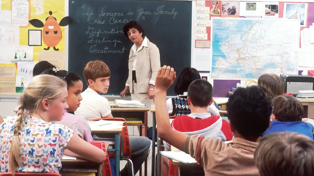 Teacher presenting a lesson in front of a chalkboard while students sit at desks and one boy raises his hand to speak