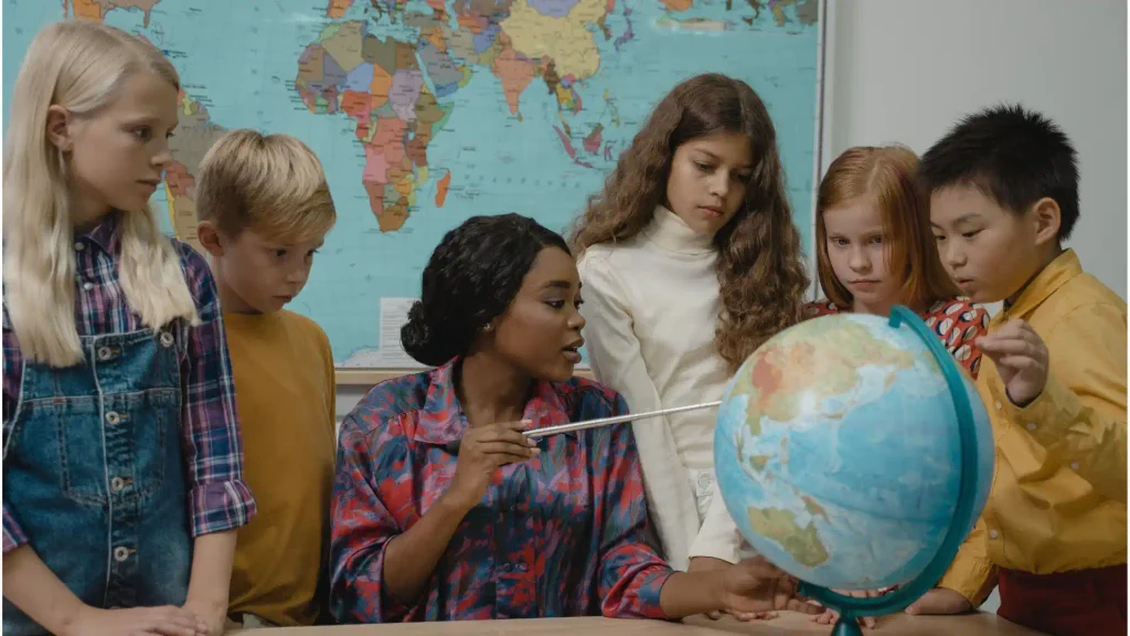 Elementary school teacher pointing at a desktop globe while explaining geography to a group of engaged young students