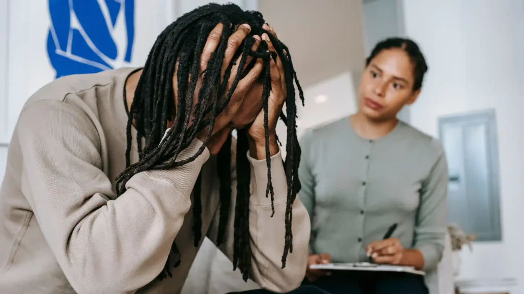 Stressed man holding his head in distress while sitting in a therapy session as a counselor takes notes on a clipboard
